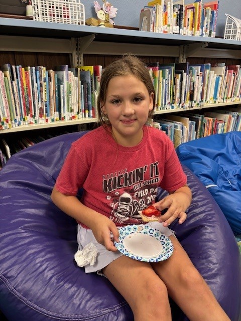 Child in red shirt on purple bean bag holding plate of food, seated in front of bookshelves in a library.